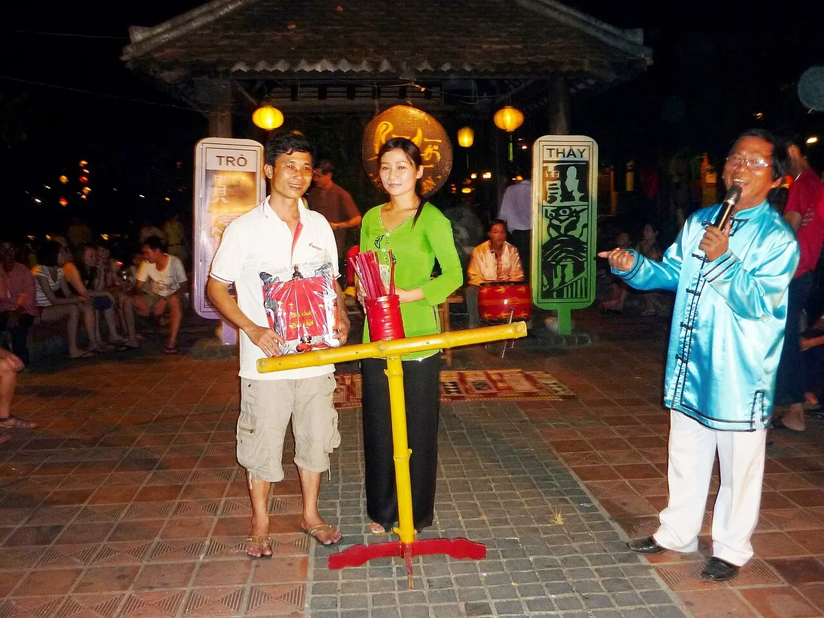 Night scene in Hoi An: a man in a light blue silk jacket sings into a microphone beside a woman and a man standing at a yellow stand, with a drum and painted boards behind them.
