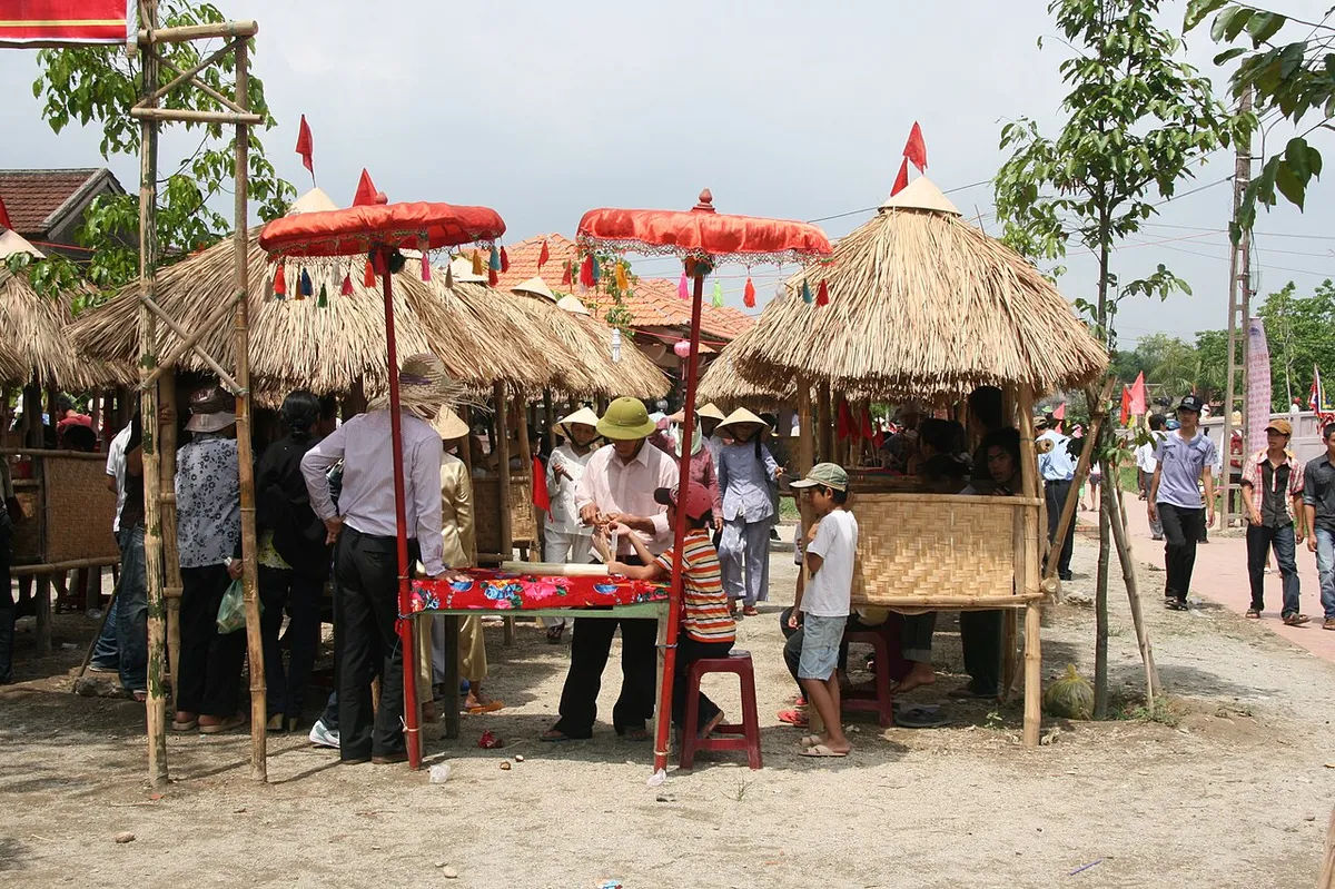 Những chòi lợp tranh dựng trên khung tre với ô nhỏ màu đỏ, mọi người đứng quanh một chiếc bàn phủ khăn hoa trên nền đất nắng.