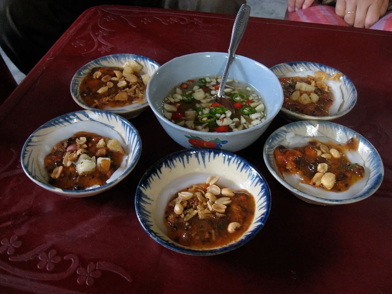 Five blue-rimmed bowls of steamed rice cakes with a savoury topping and peanuts around a bowl of fish sauce with chilli