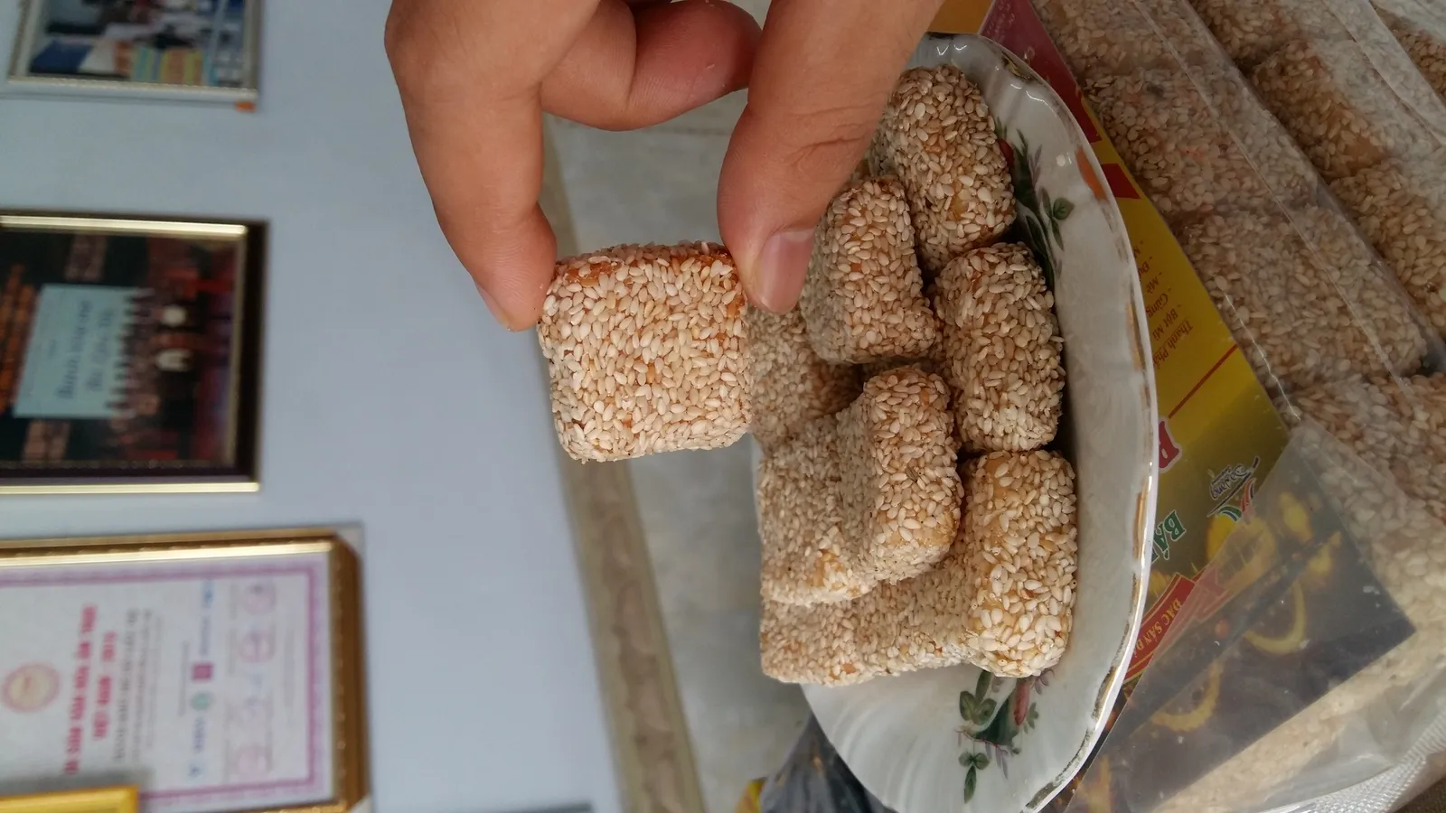 Chiếc bánh vuông phủ mè được kẹp giữa hai ngón tay phía trên đĩa bánh, phía trước là các hộp bánh.