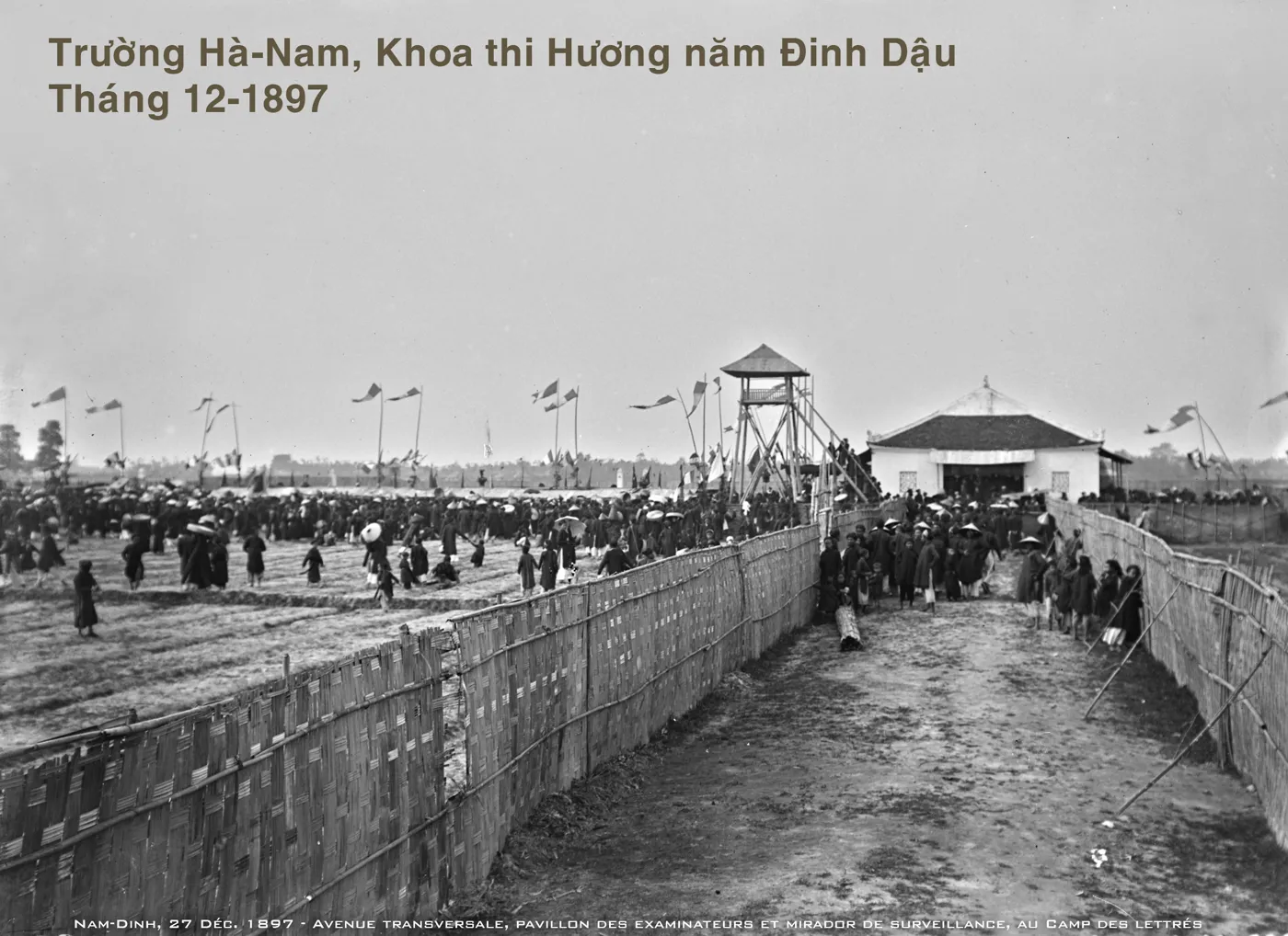 Ảnh đen trắng bãi thi rộng tháng 12 năm 1897 quây phên tre, có chòi canh, cờ và đám đông sĩ tử.
