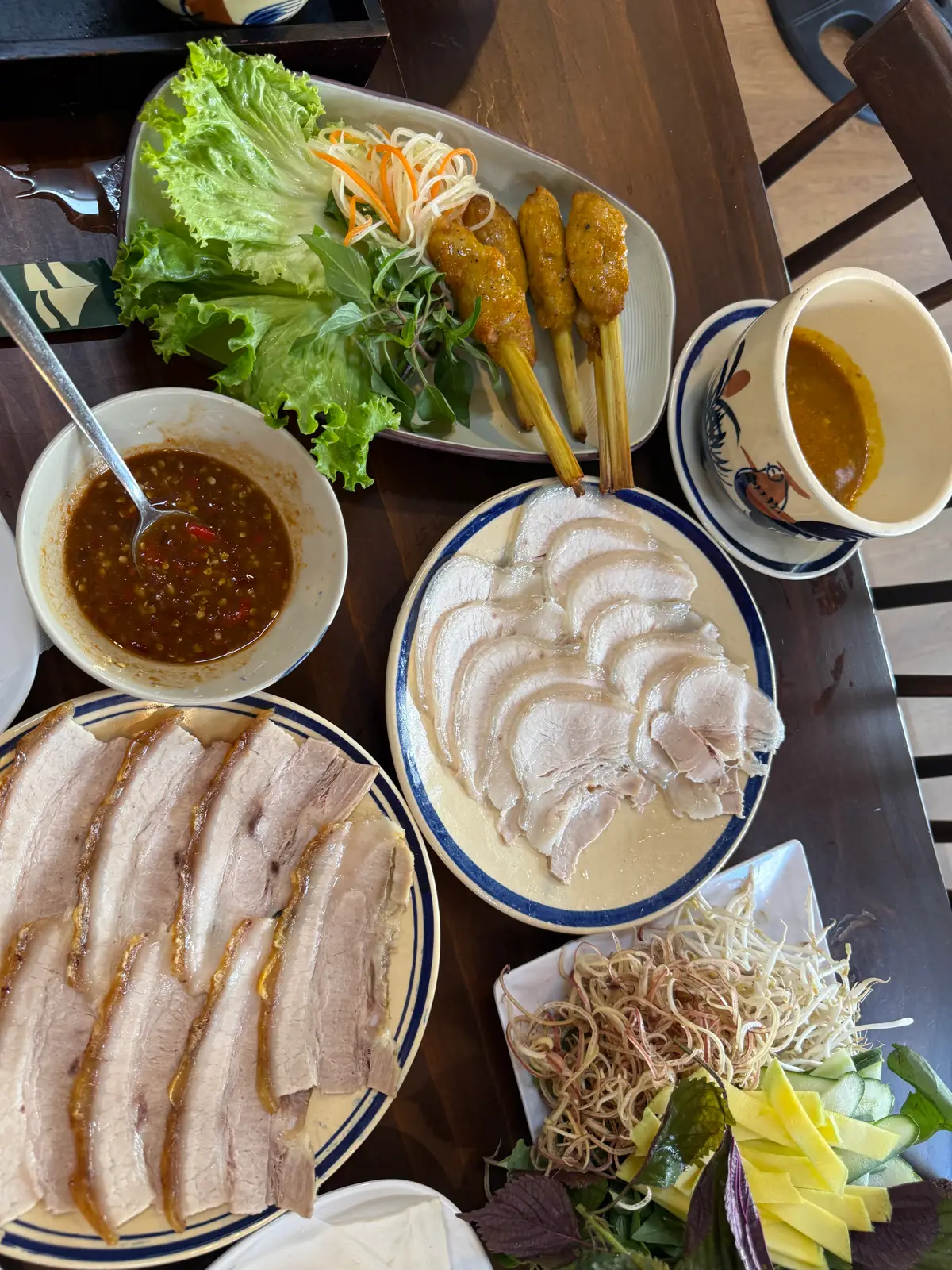 Two plates of thinly sliced boiled pork, a bowl of mam nem and herbs on a wooden table