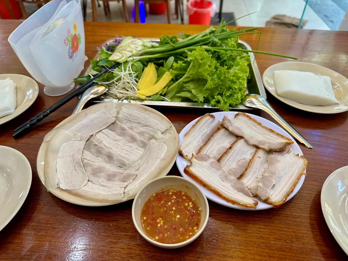 Plates of boiled belly pork and roast pork with a tray of herbs, rice paper and a red bowl of mam nem