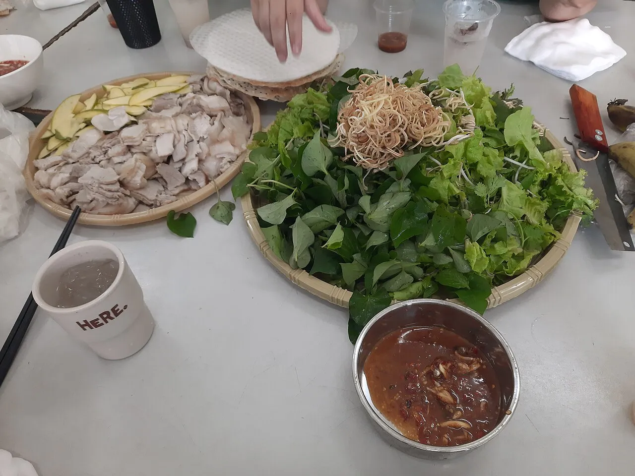 Table with a tray of sliced boiled pork and green fruit, a basket of raw herbs topped with fried noodles, a bowl of brown dipping sauce and a stack of rice paper