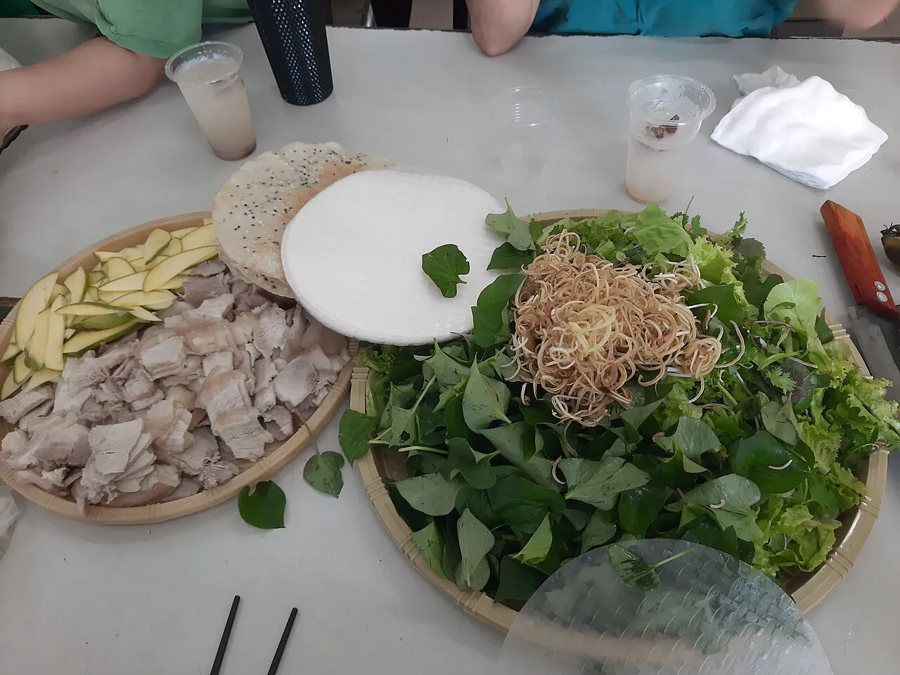 Round sheet of rice paper laid on a table beside sliced boiled pork and a basket of raw herbs