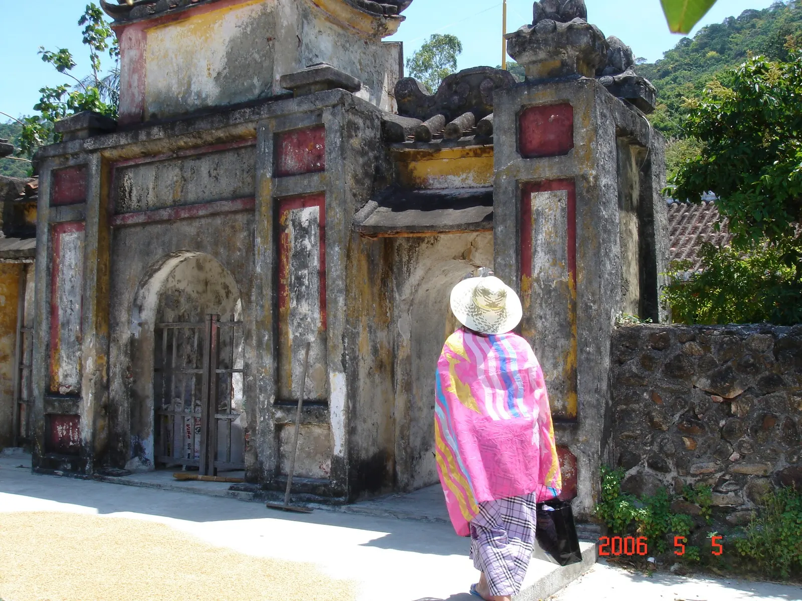 Cổng tam quan cổ của một ngôi chùa trên đảo, tường loang màu, một người đội nón khoác khăn hồng đứng phía trước.