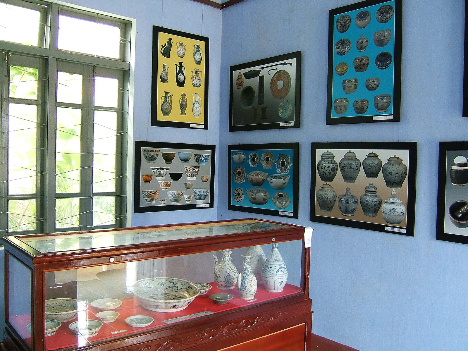 Tủ kính trưng bày bát, bình gốm hoa lam, trên tường treo các khung ảnh chụp bình, lọ, đĩa gốm.