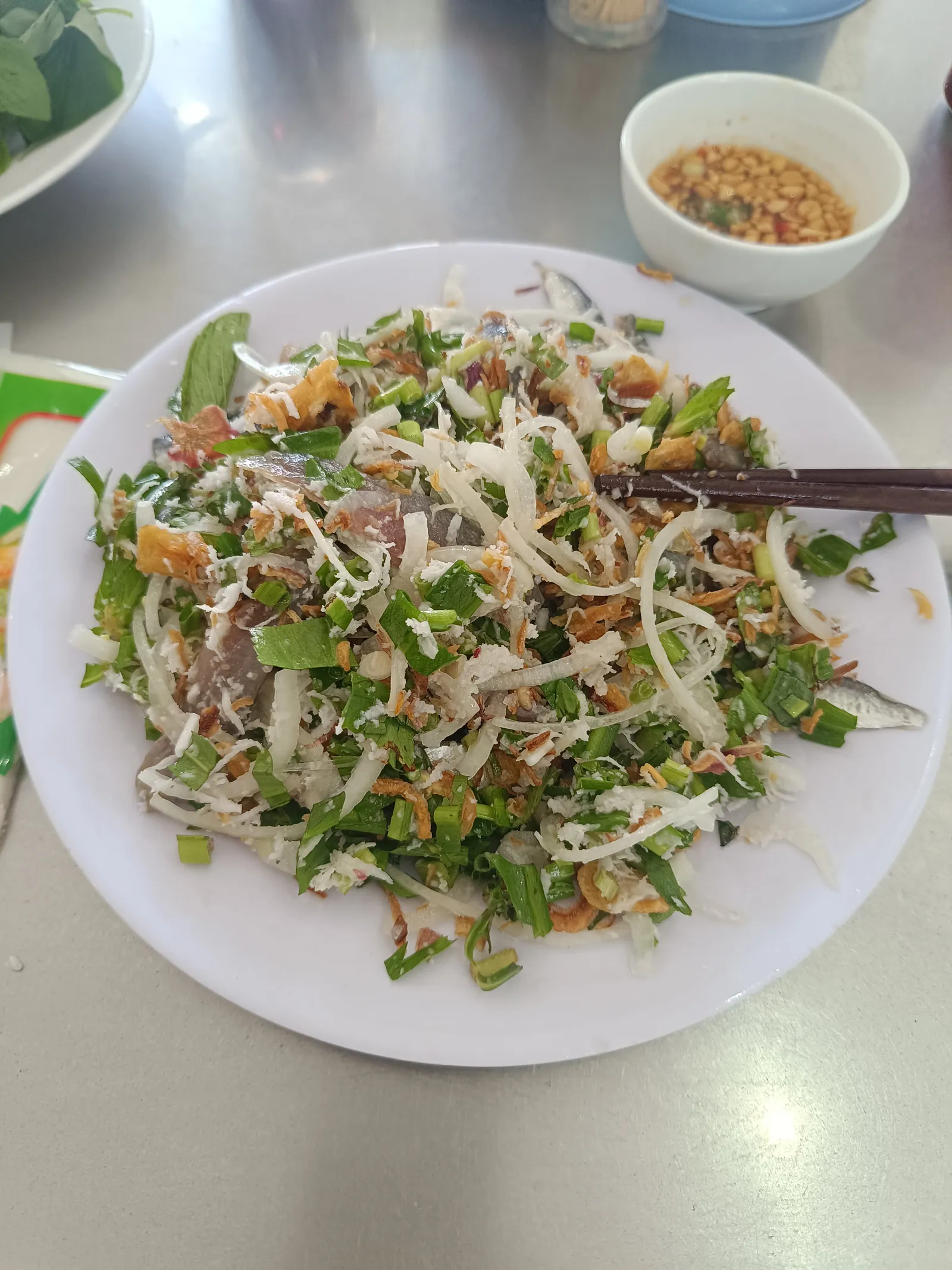 A white plate of raw herring salad mixed with green herbs, sliced onion and fried shallots, with chopsticks and a small bowl of peanut dipping sauce
