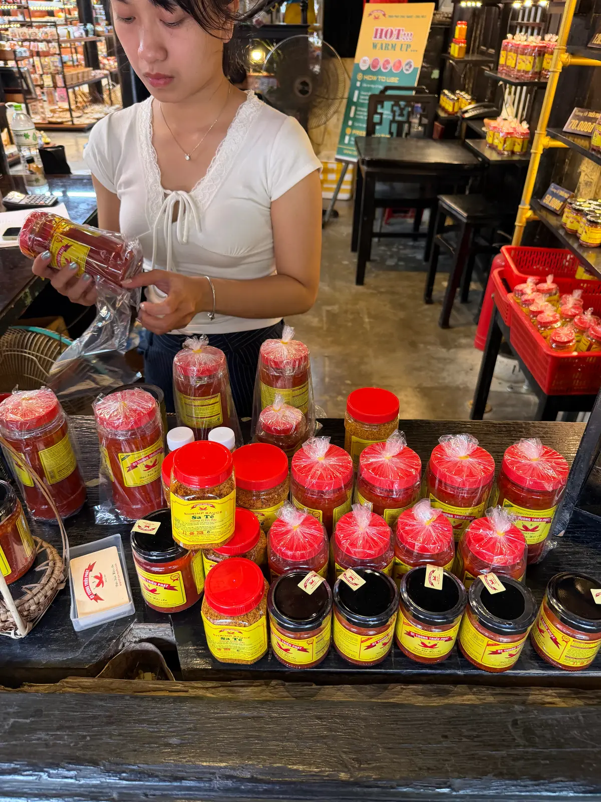 A shop assistant bagging jars of red chilli sauce with yellow labels on a counter, more jars lined up in front.