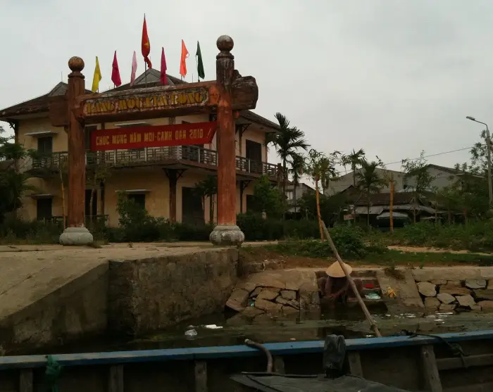 Cổng gỗ chạm của làng mộc Kim Bồng nhìn từ thuyền trên sông, phía sau là tòa nhà hai tầng màu vàng và những lá cờ.