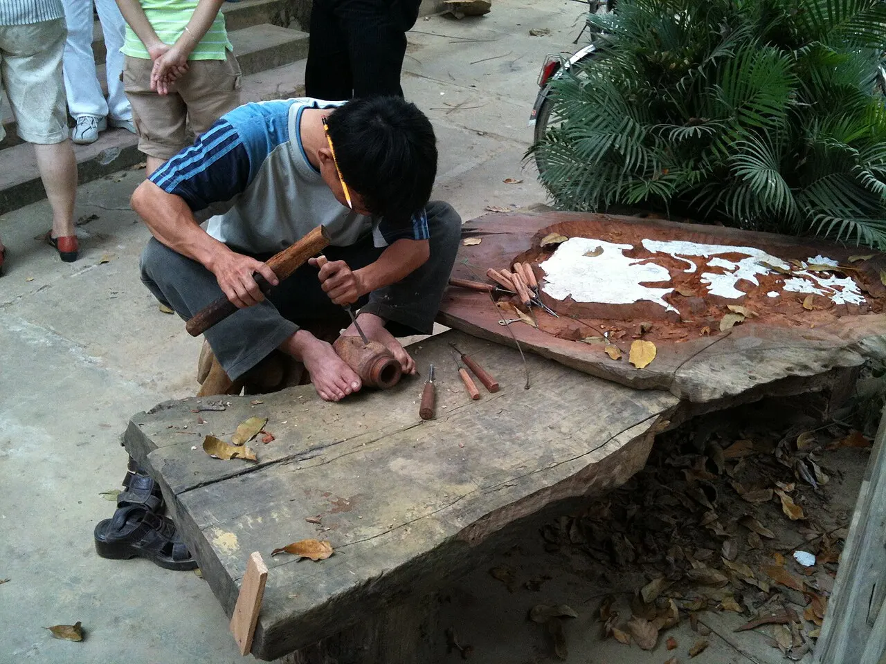 Một thợ chạm ngồi dưới đất đục một khúc gỗ, dụng cụ đục bày trên tấm ván dày bên cạnh.