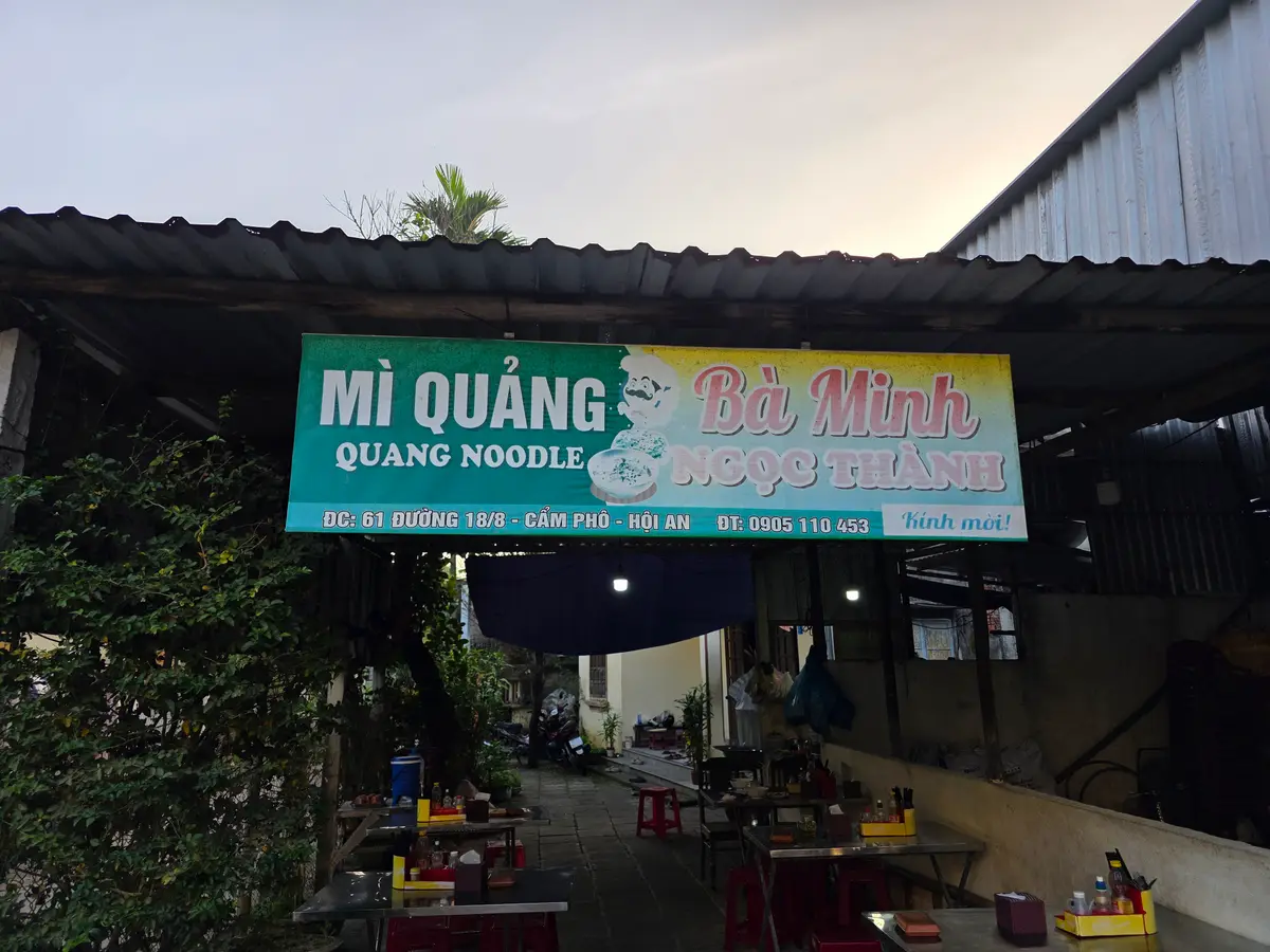 Biển hiệu Mì Quảng Bà Minh và dãy bàn ghế nhựa dưới mái che.