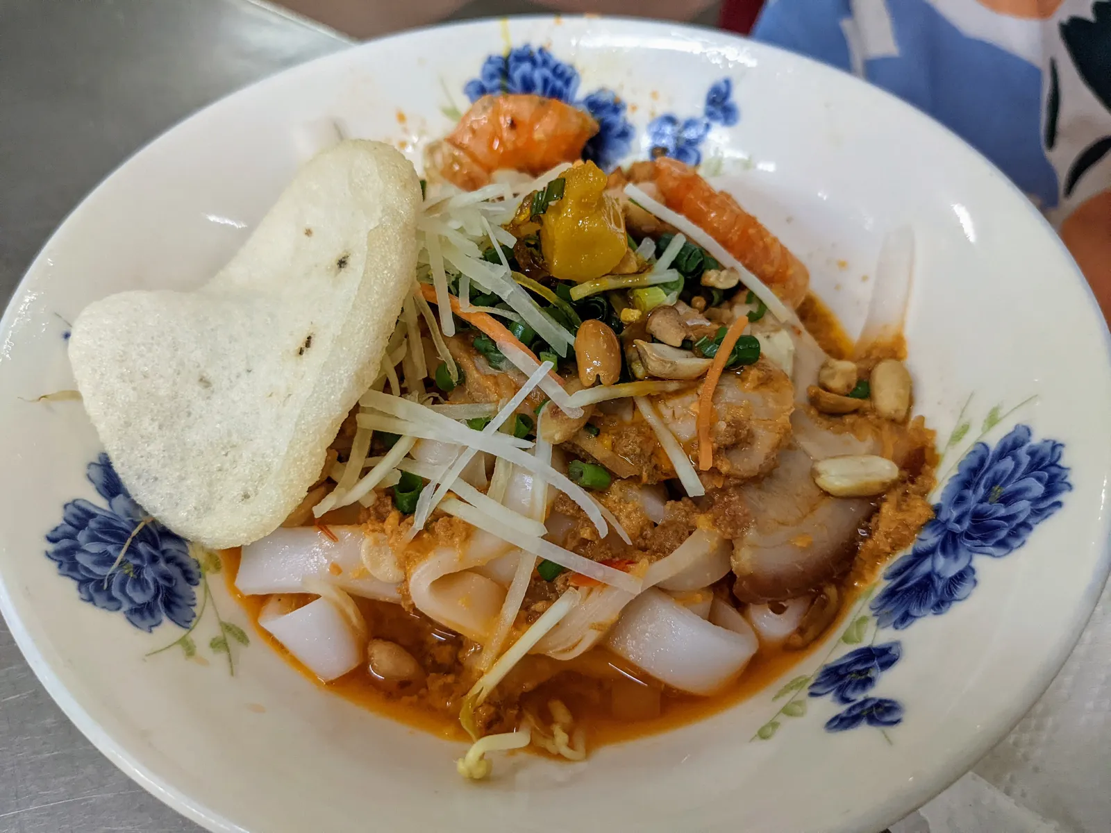 Tô Mì Quảng hoa xanh với thịt heo, tôm, đậu phộng, rau thái nhỏ và miếng bánh tráng hình trái tim tựa vào thành tô.
