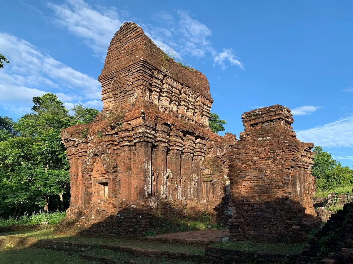 Two weathered brick towers under a blue sky, one still with many carved tiers and one partly collapsed.