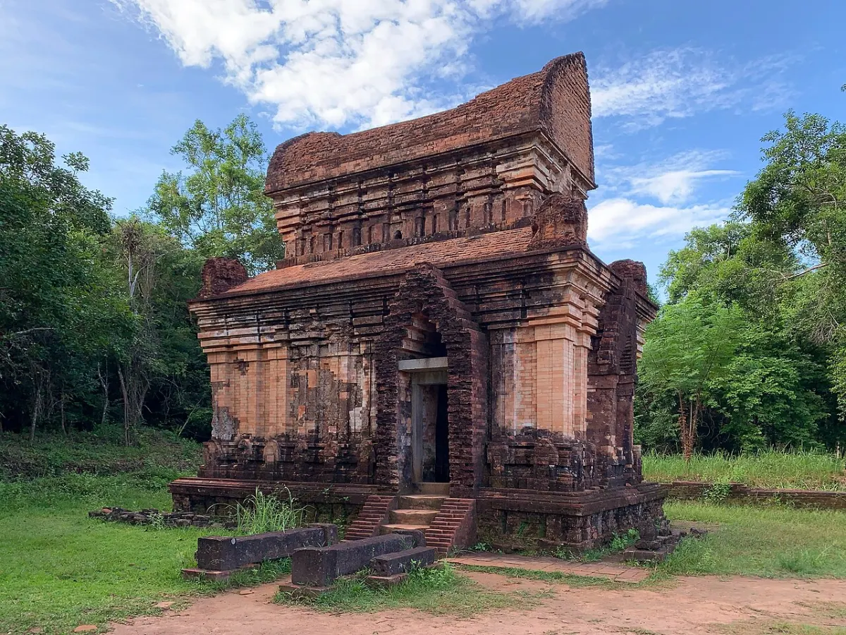 A single brick tower, kalan E7, with steps in front of the door and green grass around its base.